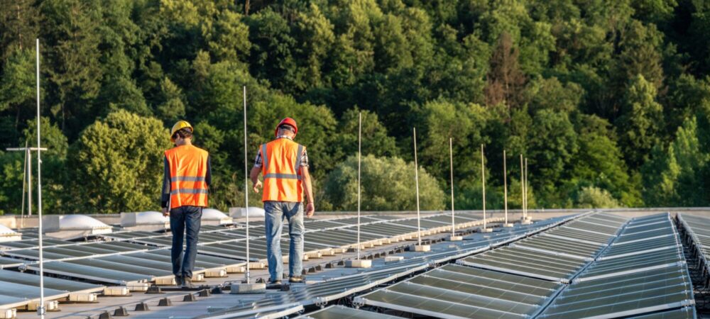 Two engineers in safety vests and helmets actively engage with a digital tablet while optimizing a solar power installation atop a spacious rooftop.